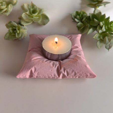 Pillow Shaped Tea Light Holder, Pink in Colour, Sat on a white surface with succulents in the background.