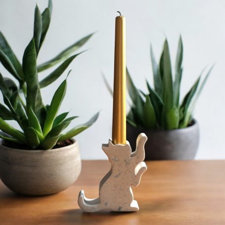 White cande holder shaped like a begging cat holding a gold candle. Resting on a wooden table with 2 plants in the background.